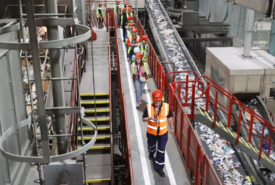 An image of people on a tour of a recycling facility. The people are wearing orange vests, red helmets and are walking along a path overlooking conveyor belts. 