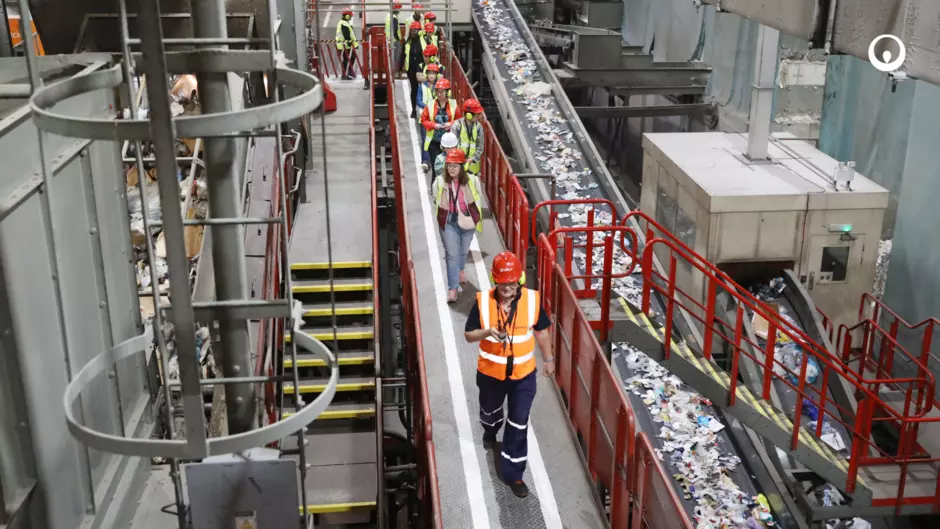 An image of people on a tour of a recycling facility. The people are wearing orange vests, red helmets and are walking along a path overlooking conveyor belts. 